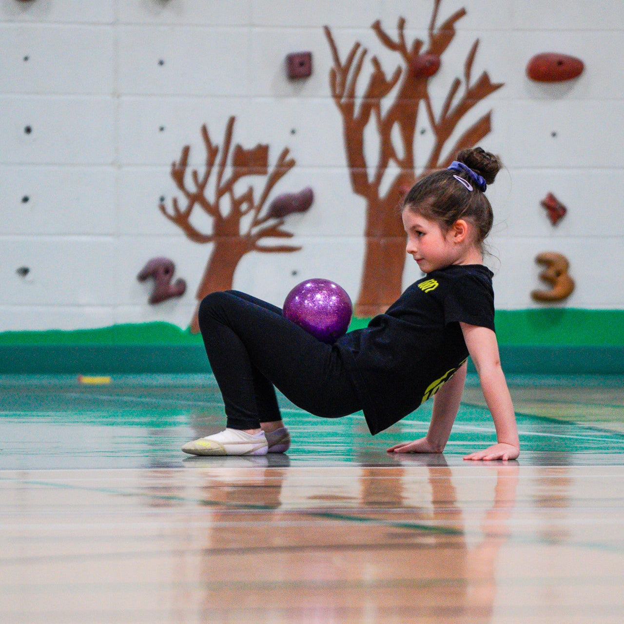 Young rhythmic gymnast holding purple ball on stomach in crab warm up