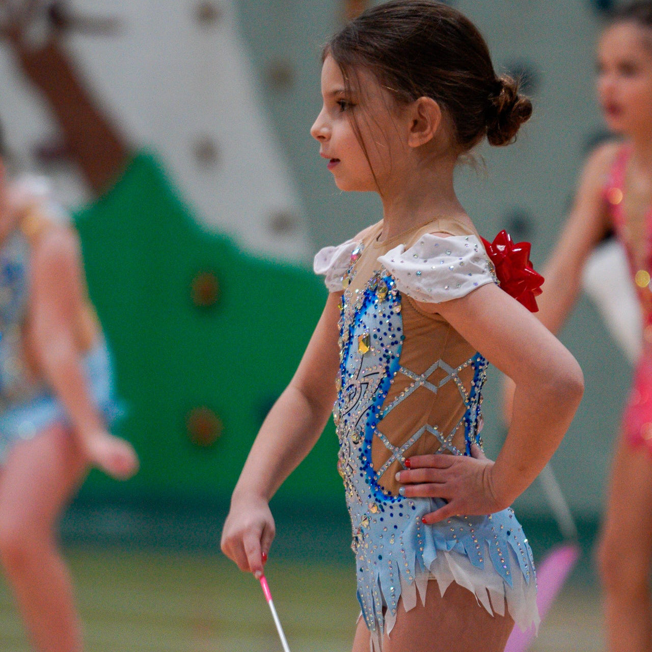 Young girl in a rhythmic gymnastics outfit with a ribbon, standing on a sports floor.