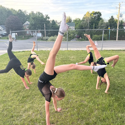 Five young gymnasts in black gemmez sportswear performing acrobatic moves on a grassy field.