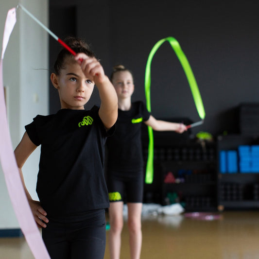 Two children practicing with colorful gymnastics ribbons in a indoor gym, one in pink and one in green.